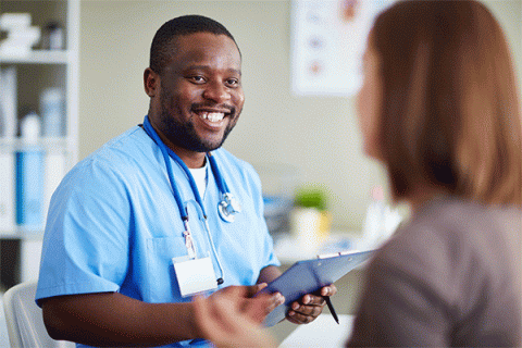 Black male with short hair, beard and mustache wearing blue nursing scrubs and a nametag and holding a clipboard smiles and talks to a female patient with her back to the camera in a medical office setting.   